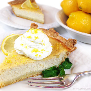 A slice of this authentic Italian ricotta pie recipe on a white plate with whipped cream, lemon zest, and fresh mint, as well as Meyer lemons and another slice of this ricotta cheesecake in the background