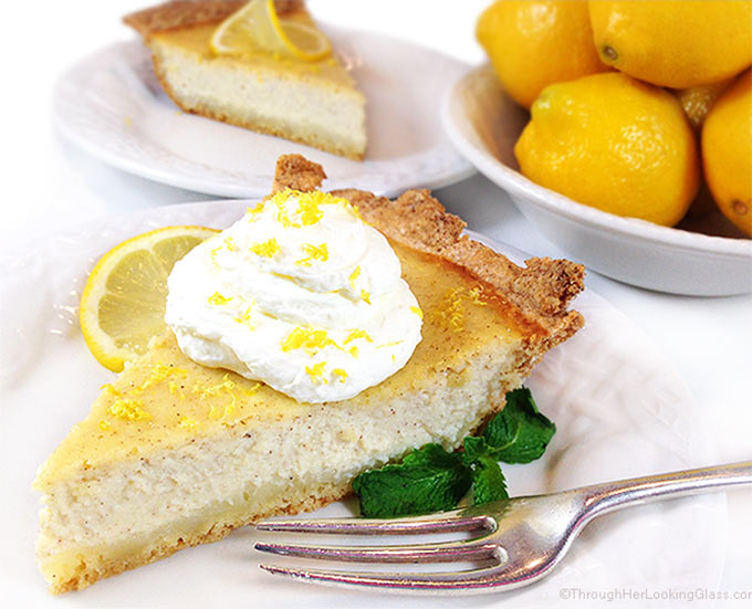 A slice of this authentic Italian ricotta pie recipe on a white plate with whipped cream, lemon zest, and fresh mint, as well as Meyer lemons and another slice of this ricotta cheesecake in the background